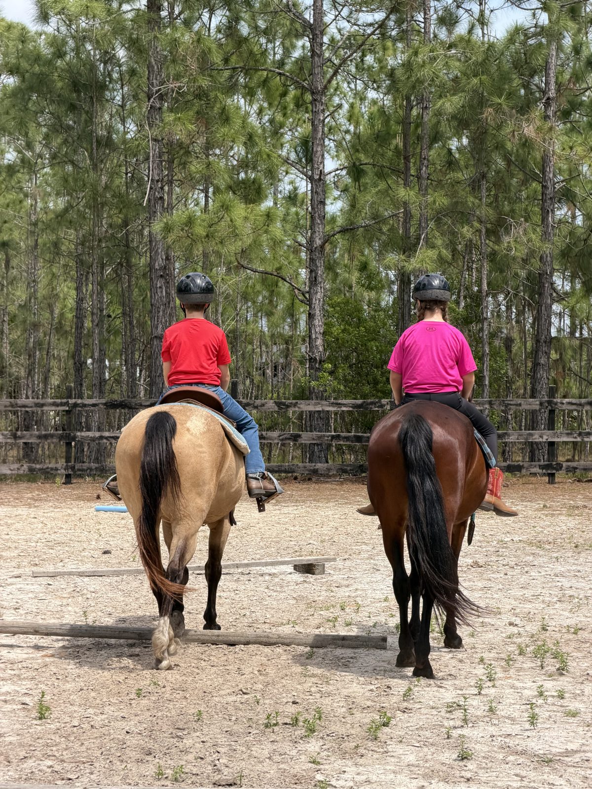Two children ride side by side in the sandy arena